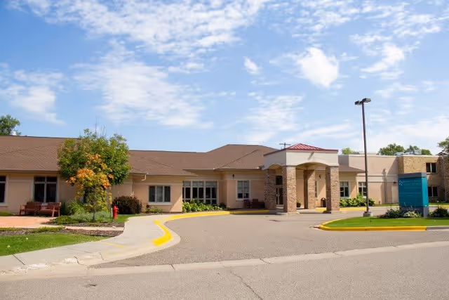 Exterior view of Benedictine Living Community-New Brighton building under a partly cloudy sky, featuring a driveway with a covered entrance, landscaped greenery, and a sign near the entrance.