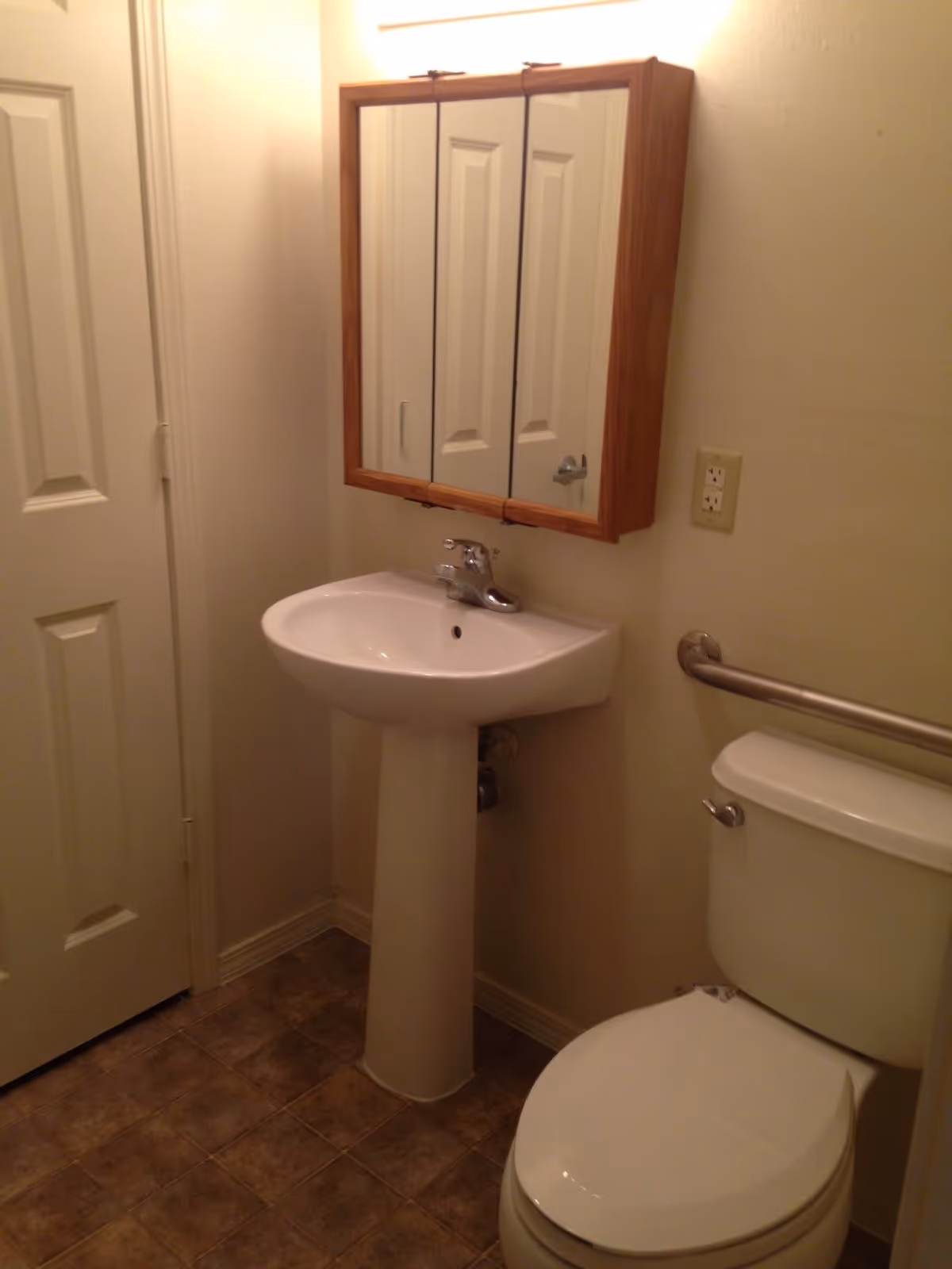 A small bathroom with a white pedestal sink, a toilet with a grab bar on the wall beside it, a wooden framed mirrored medicine cabinet above the sink, and a closed door to the left. The floor has brown tiles and the walls are painted light beige.