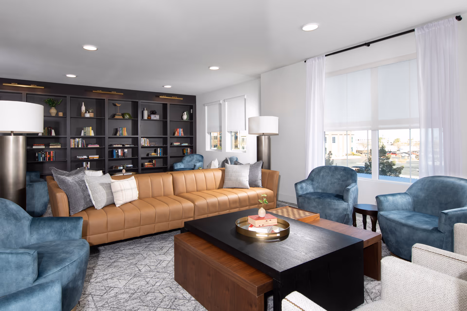 A modern living room with a long tan leather sofa adorned with multiple pillows, surrounded by blue upholstered armchairs and beige chairs. A large black and wooden coffee table sits in the center with a decorative tray and books. The back wall features a dark built-in bookshelf filled with books and decorative items. Large windows with white curtains allow natural light to fill the room.