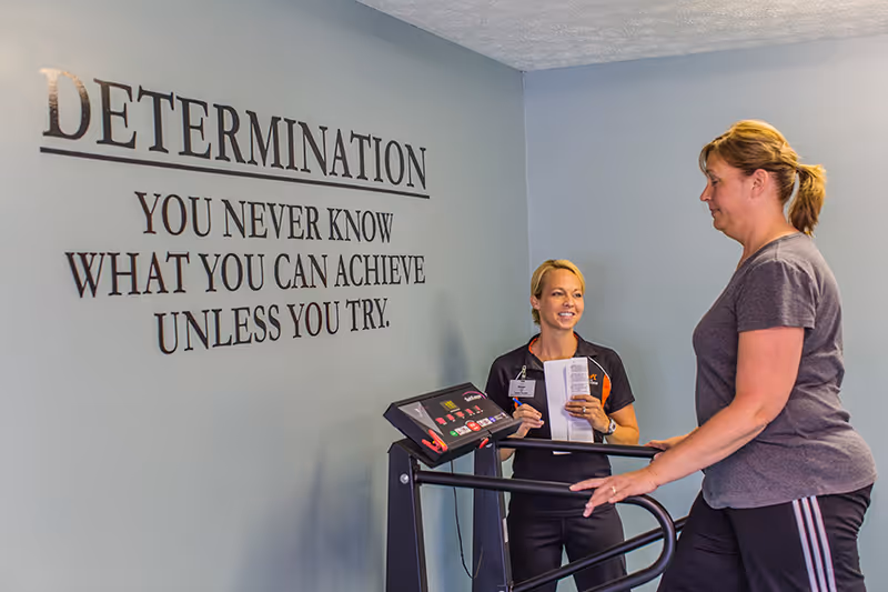 A woman in workout clothes is walking on a treadmill while another woman, possibly a trainer or therapist, stands beside her holding a clipboard and smiling. On the wall behind them is a motivational quote that reads: 'DETERMINATION - YOU NEVER KNOW WHAT YOU CAN ACHIEVE UNLESS YOU TRY.'