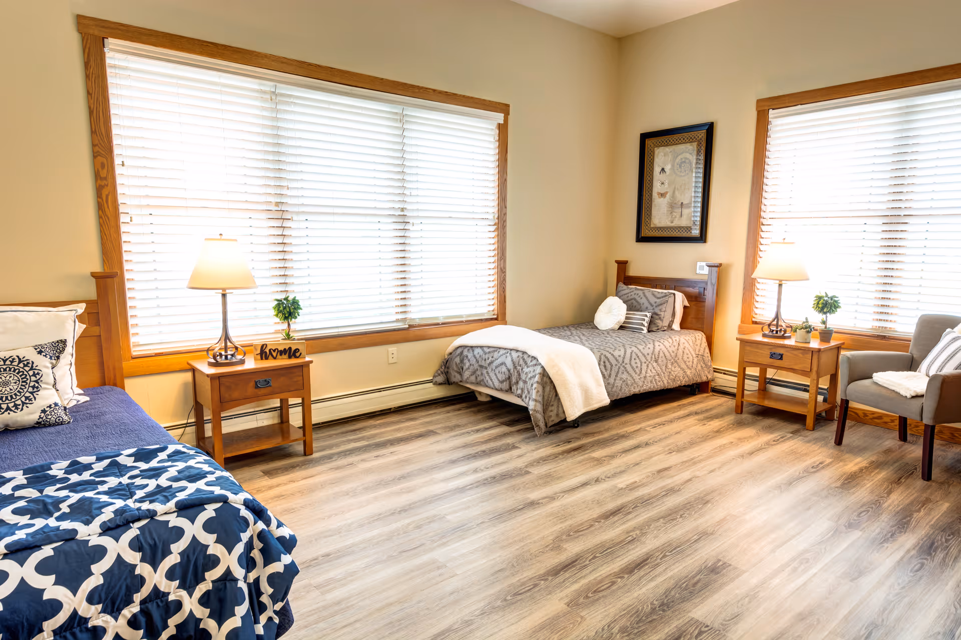 A bright and cozy bedroom with two single beds, each with patterned bedding and pillows. Between the beds are wooden nightstands with table lamps and small potted plants. Large windows with white blinds allow natural light to fill the room. There is a framed picture on the wall and a comfortable armchair with a throw blanket near one window. The floor is wood laminate.