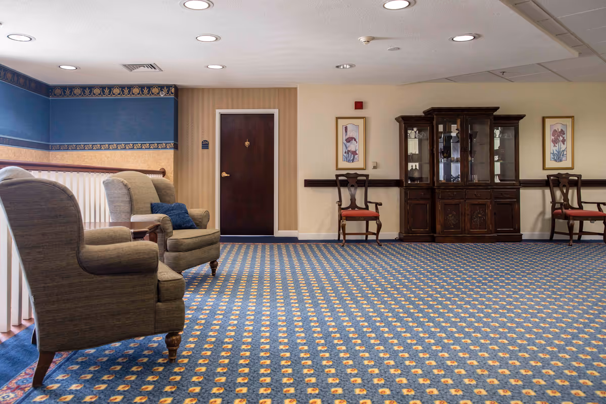 A spacious interior common area with patterned blue carpet, two upholstered armchairs with a small table between them on the left, a dark wooden door in the center, and a dark wooden china cabinet flanked by two wooden chairs with red cushions on the right. The walls are decorated with framed floral artwork and have a beige and blue color scheme with a decorative border near the ceiling.