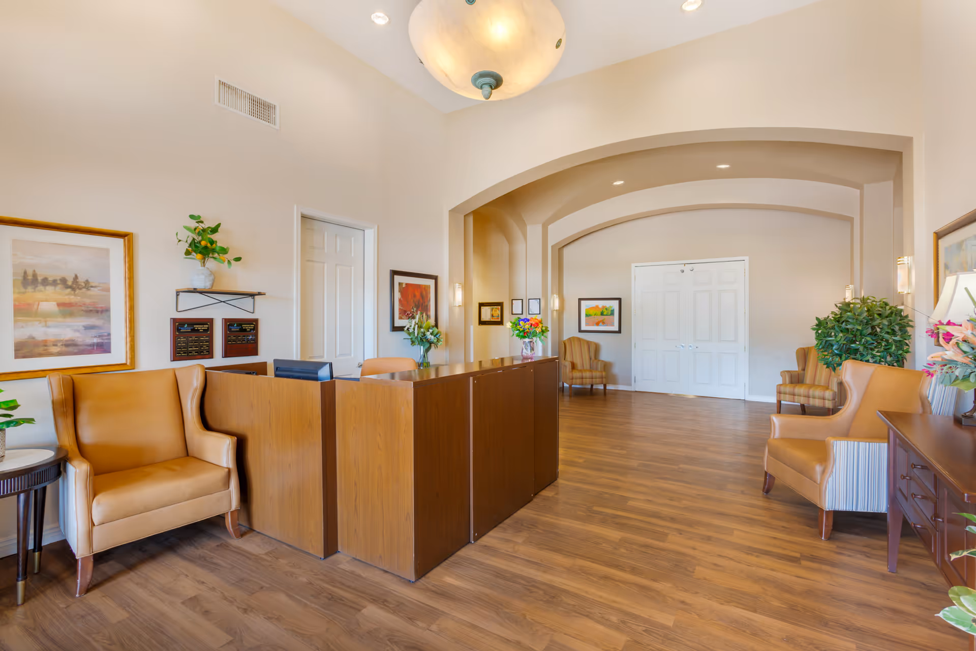 Reception desk and seating area in a bright senior living facility lobby with chairs, artwork, and plants.