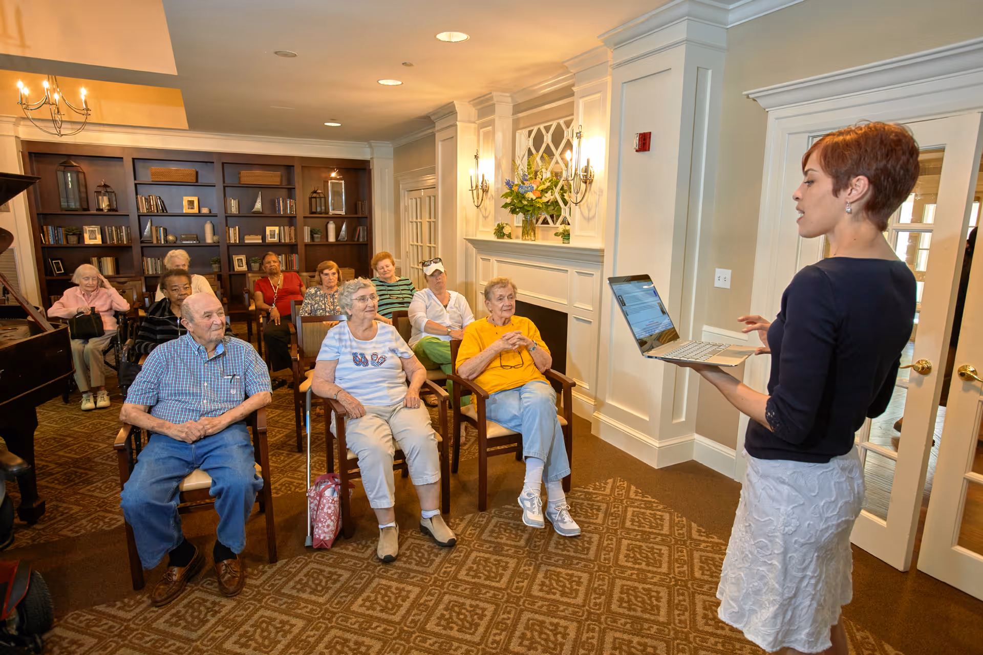 A group of elderly people seated in a room with bookshelves and a fireplace, attentively listening to a woman standing and holding a laptop, possibly giving a presentation or leading a discussion.