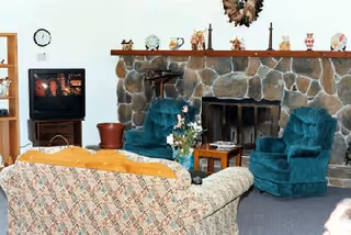 Cozy living room with a stone fireplace and mantle, two teal armchairs, a patterned sofa, TV on a stand, bookshelf, and decorative items.