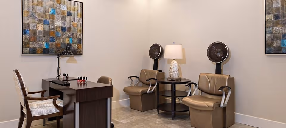 A salon area with two brown leather chairs under hair dryers, a small round table with a lamp between them, a dark wooden manicure table with nail polish bottles and a lamp, and a cushioned armchair. The walls are light-colored with two abstract paintings.
