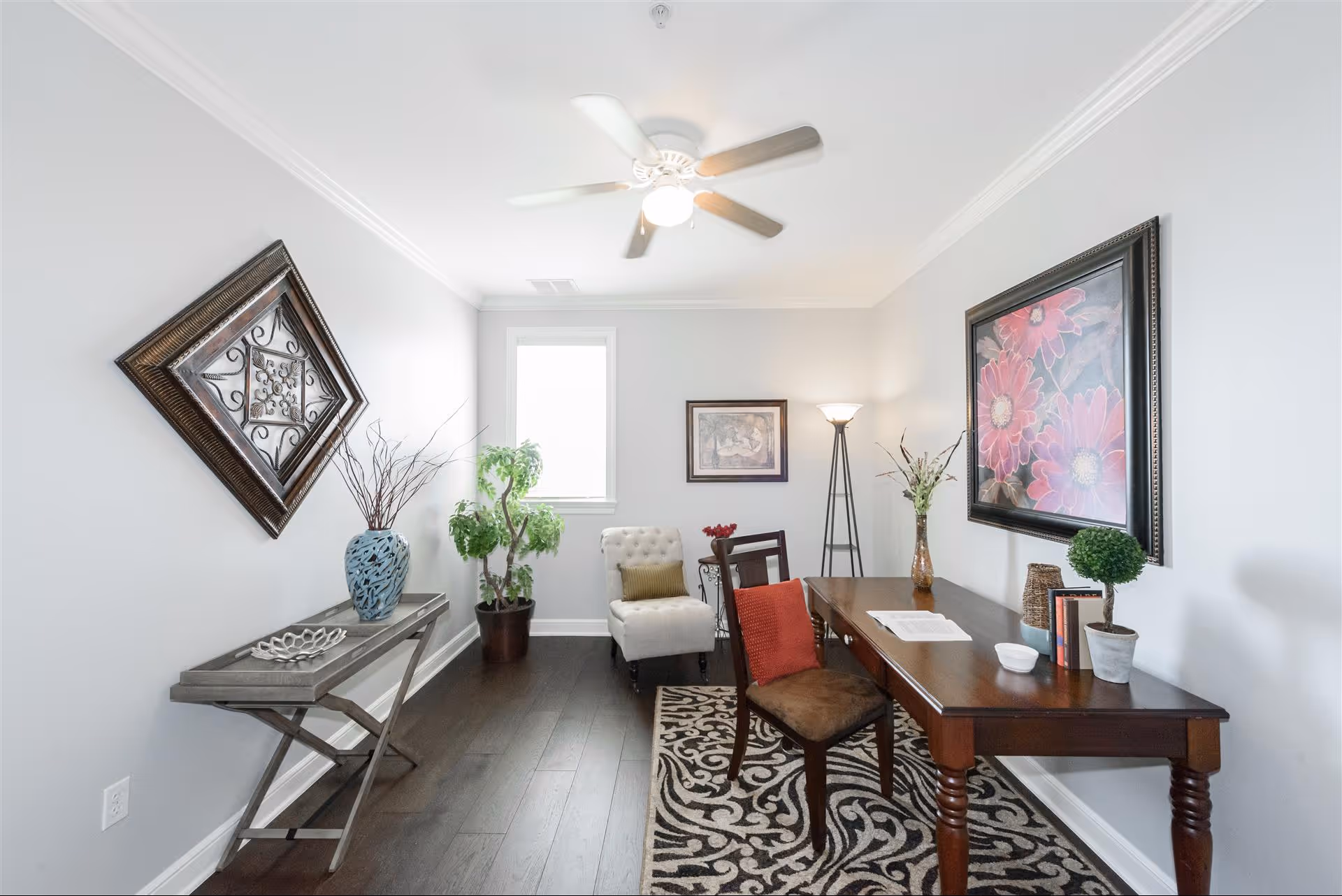 Bright home office-style sitting room with a wooden desk, chairs, wall art, plants, and a ceiling fan.