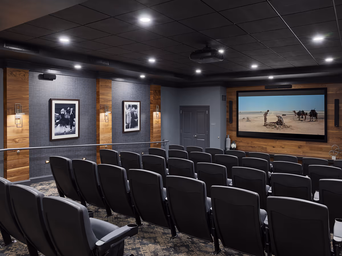 Private theater room with rows of gray seats facing a large screen showing a desert scene and framed photos on wood-paneled walls.