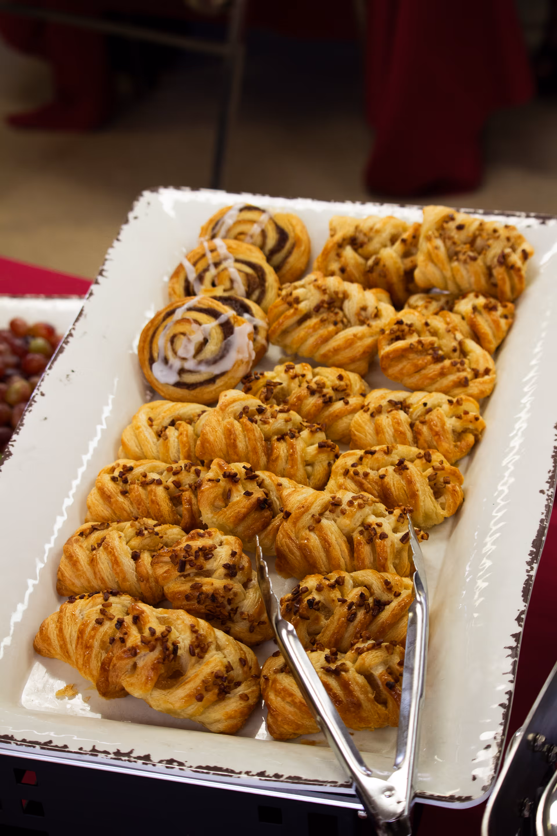 Tray of assorted pastries and cinnamon rolls on a white serving platter with metal tongs.
