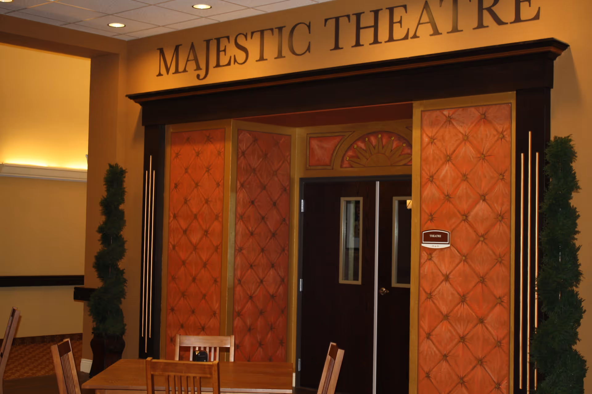 Entrance to the Majestic Theatre with double doors framed by orange padded panels and gold trim, flanked by two tall green plants. A wooden table with chairs is in the foreground.