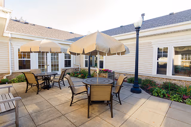 Sunlit courtyard patio with round tables, umbrellas, and chairs beside a single-story building.