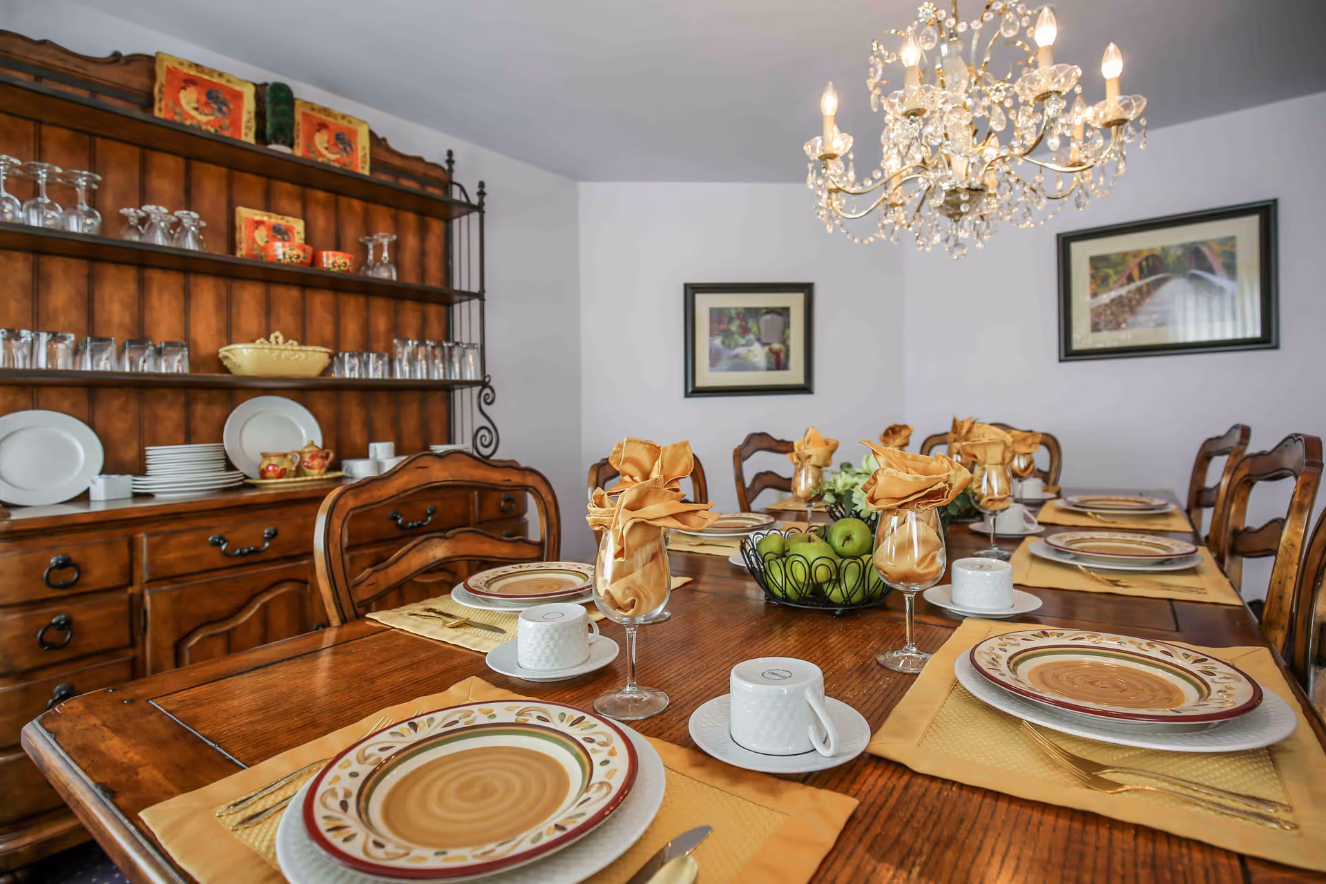 A dining room with a wooden table set for six people. The table has plates, cups, wine glasses with folded napkins, and a centerpiece with green apples. There is a wooden hutch in the background holding plates, glasses, and decorative items. A chandelier hangs above the table, and two framed pictures are on the wall.