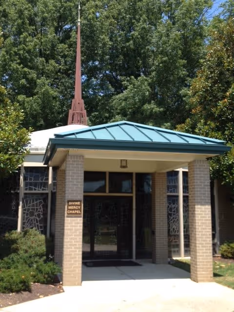 Entrance to Divine Mercy Chapel featuring a covered porch with brick pillars and a teal metal roof, surrounded by trees and shrubs.