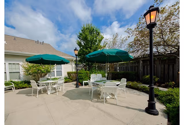 Outdoor patio area with two round tables, each shaded by a green umbrella, surrounded by white chairs. The patio is paved and bordered by greenery, with a wooden fence and a building with beige siding in the background. Two black lamp posts with lit lanterns are also visible.