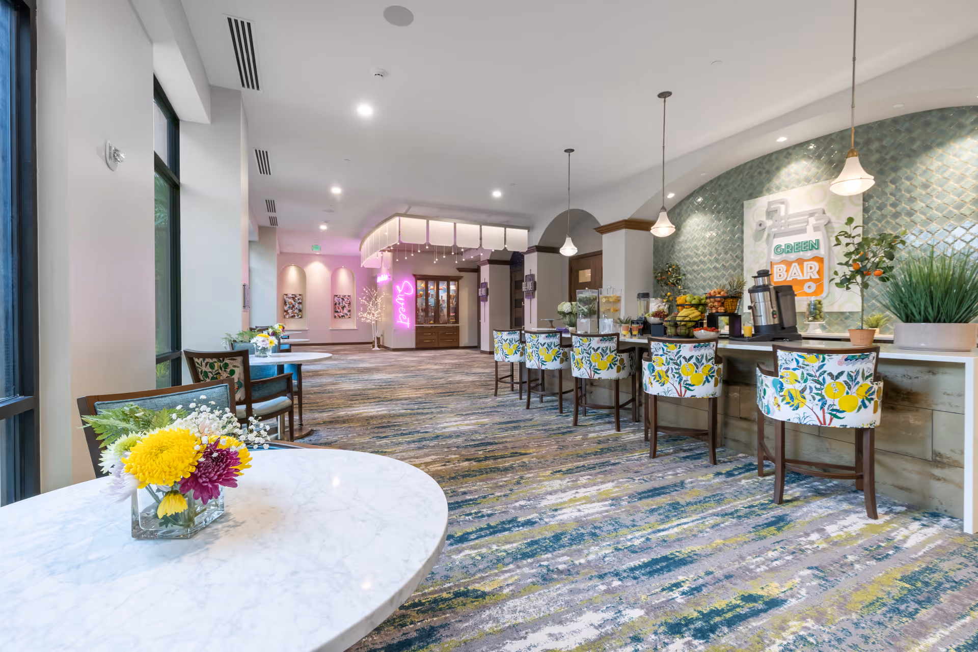 Bright communal lounge and bar area with floral-upholstered bar stools, marble tables with flower arrangements, and a counter labeled 'Green Bar'.