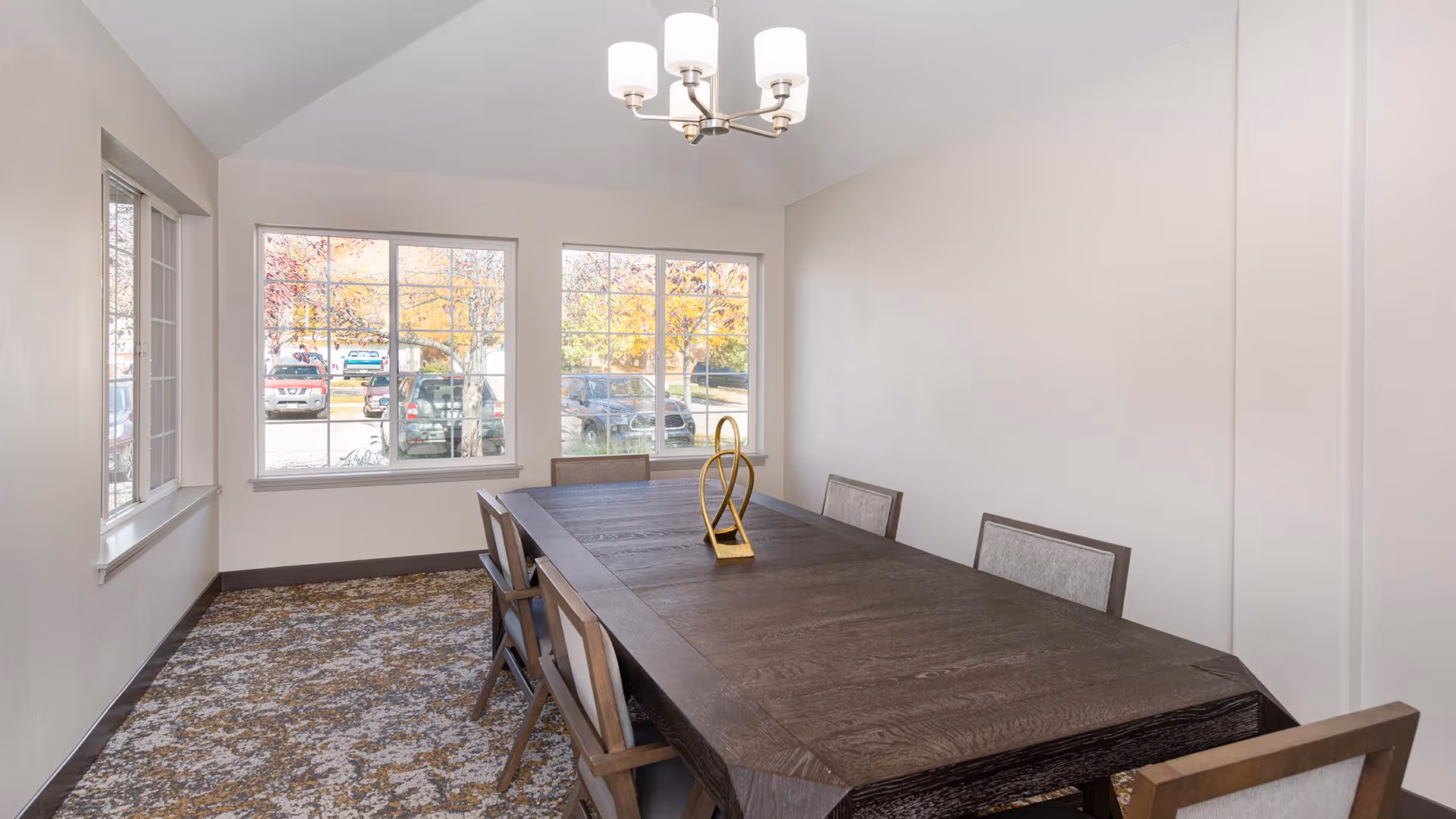 Bright dining room with a long dark wood table and chairs, large windows showing parked cars outside, and a ceiling chandelier.
