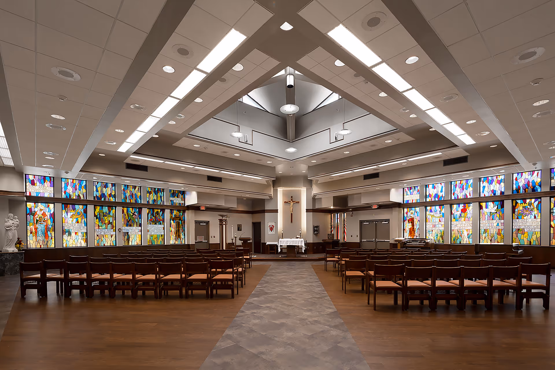 Spacious chapel interior with rows of wooden chairs, colorful stained glass windows, and an altar topped by a crucifix.