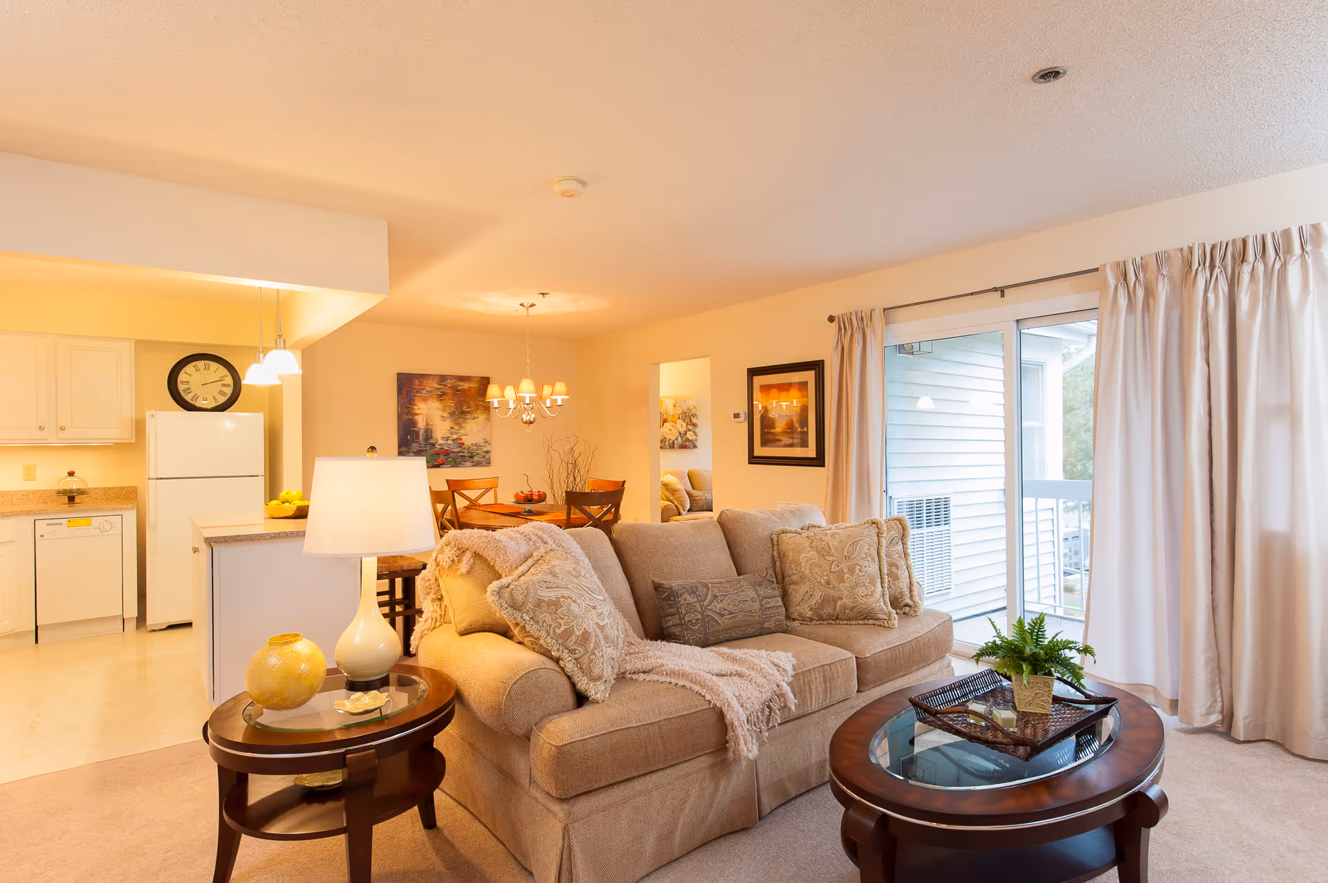 A cozy living room area with a beige sofa adorned with patterned cushions and a throw blanket. In front of the sofa is a round wooden coffee table with a glass top and a small potted plant. To the left, there is a side table with a lamp and decorative items. Behind the sofa, an open dining area with a wooden dining table and chairs is visible, along with a kitchen area featuring white cabinets, a refrigerator, and a clock on the wall. Large sliding glass doors with curtains lead to an outdoor balcony.