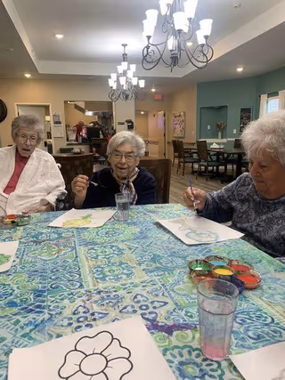 Three elderly women sitting around a table covered with a blue patterned tablecloth, engaged in painting activities with watercolor paints and brushes. The room is well-lit with chandeliers, and there are tables and chairs in the background, suggesting a communal indoor space.