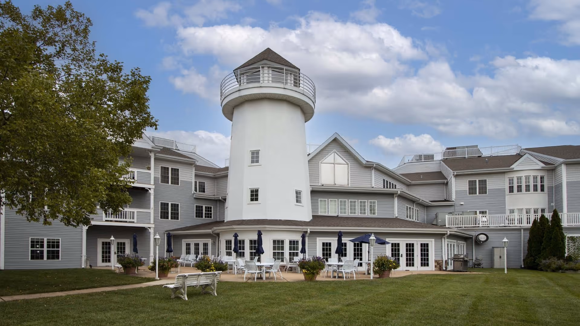 Front exterior of a gray multi-story assisted living building with a central white lighthouse tower, outdoor seating, and a grassy lawn.