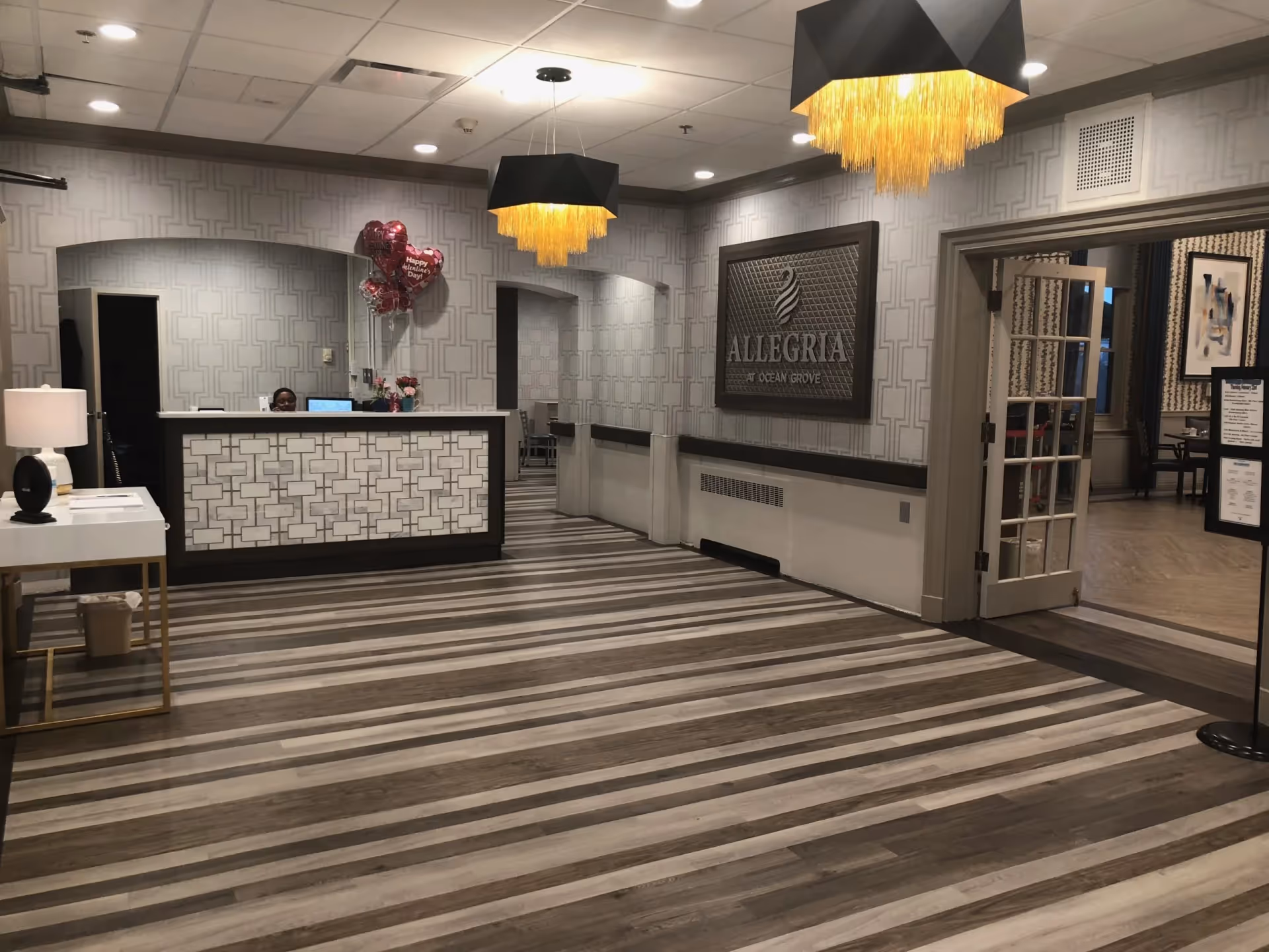 Reception area of a senior living facility with a front desk, a lamp on a side table, two modern chandeliers with yellow fringe, patterned wallpaper, and a sign on the wall that reads 'ALLEGRIA at OCEAN GROVE'. There are balloons behind the desk and an open doorway leading to another room with tables and chairs.