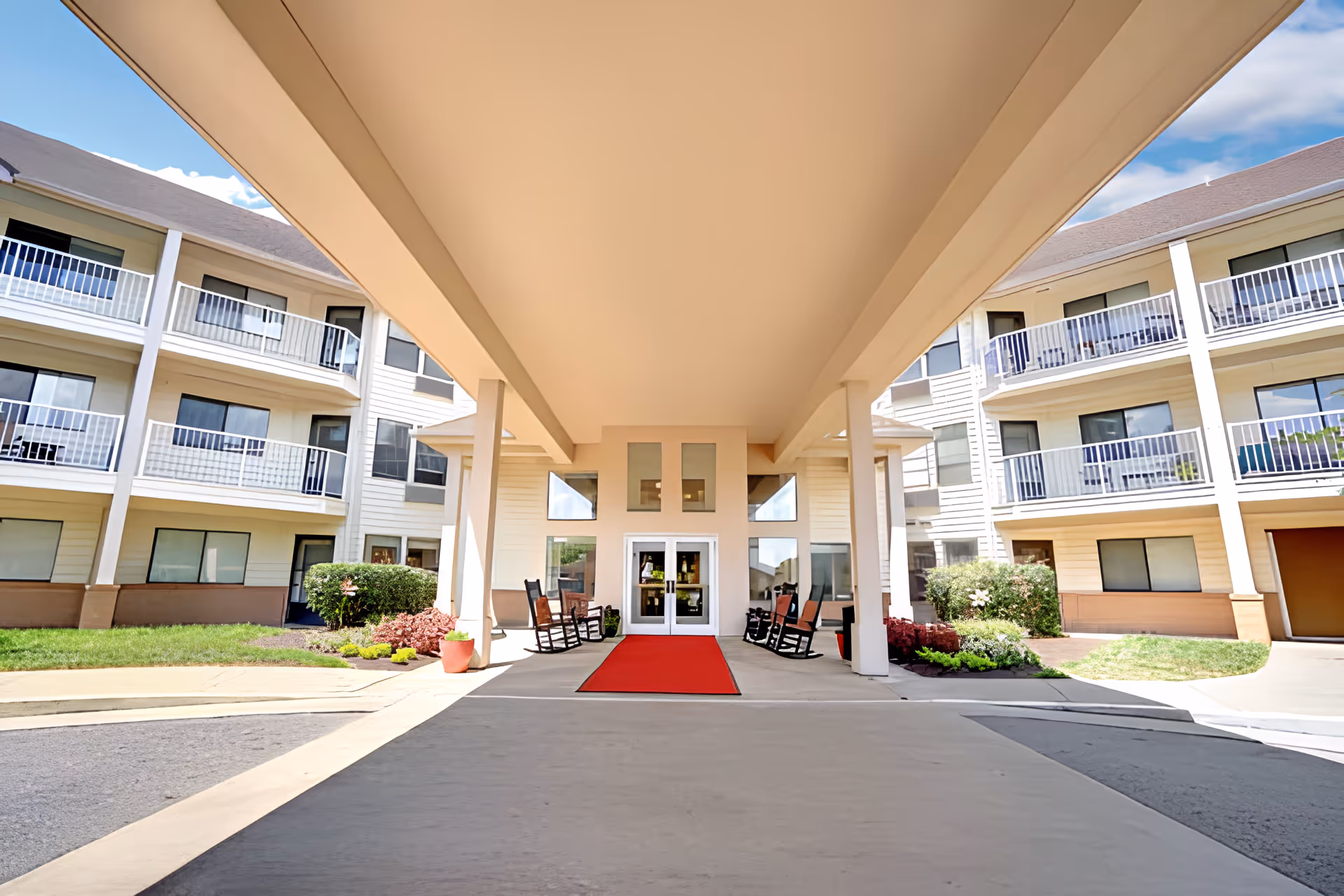 Covered entrance and portico of a multi-story senior living building with balconies, rocking chairs, and a red carpet leading to glass doors.
