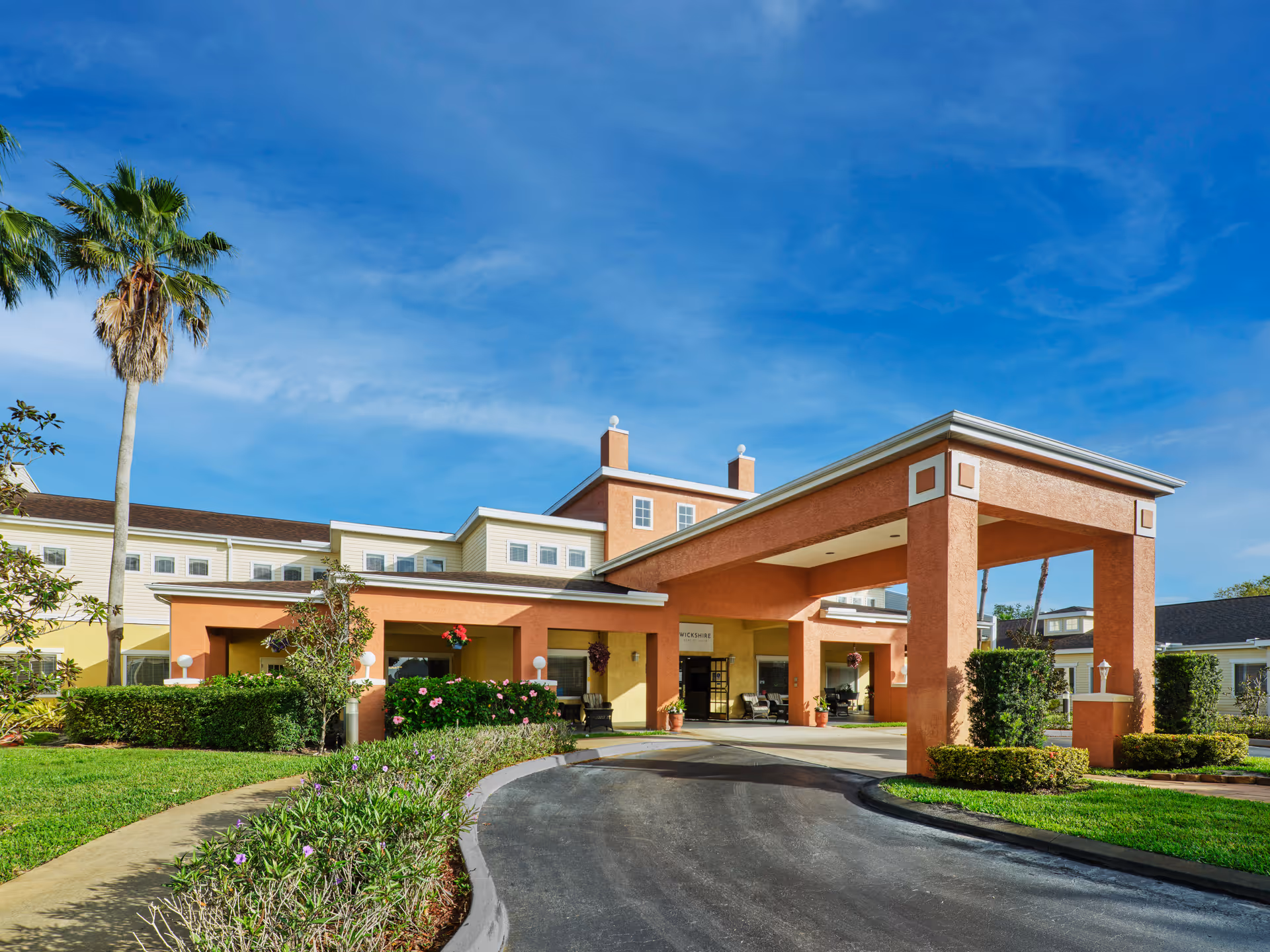 Front entrance of a salmon-colored assisted living building with a covered porte-cochère, palm trees and landscaped driveway under a blue sky.
