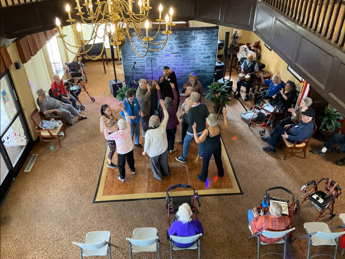 Overhead view of residents dancing on a small wooden dance floor while others sit around in a senior living facility common room.