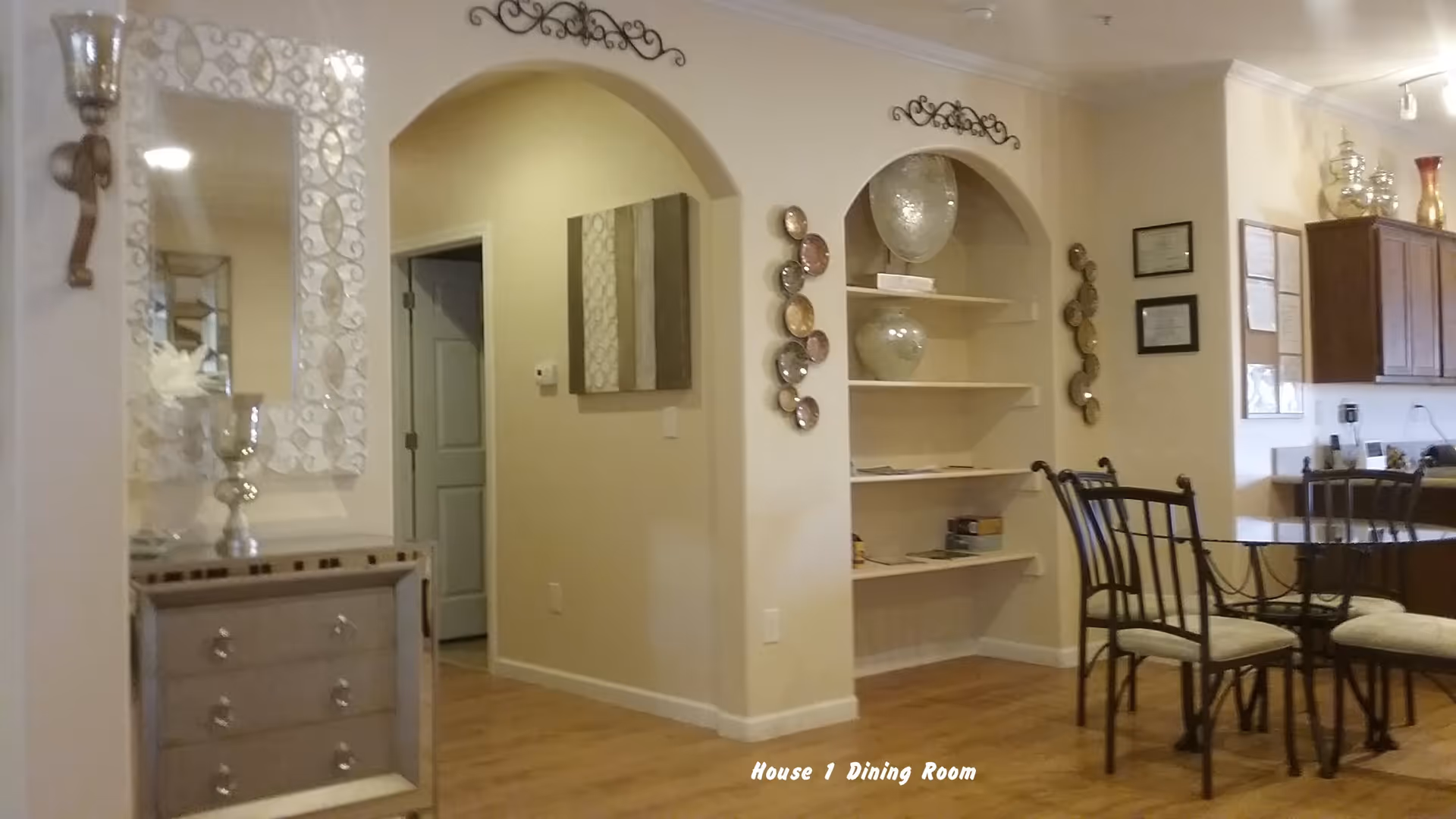 Interior view of a dining room area with a glass-top dining table and four chairs with cushioned seats. The room features light-colored walls, wooden flooring, decorative wall shelves with vases, framed certificates on the wall, and a small mirrored chest of drawers with decorative candle holders. There are two arched wall niches and decorative wall art above them.