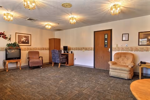 A common area in a senior living facility featuring several upholstered chairs, a small wooden table with a television, a computer desk with a computer, framed artwork on the walls, and a closed wooden door. The room has carpeted flooring and ceiling lights.