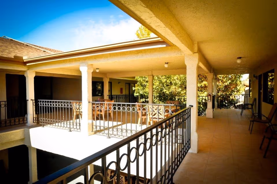 Covered outdoor balcony area with metal railing, several chairs and tables, beige tiled floor, and cream-colored columns and ceiling. Trees and blue sky are visible in the background.