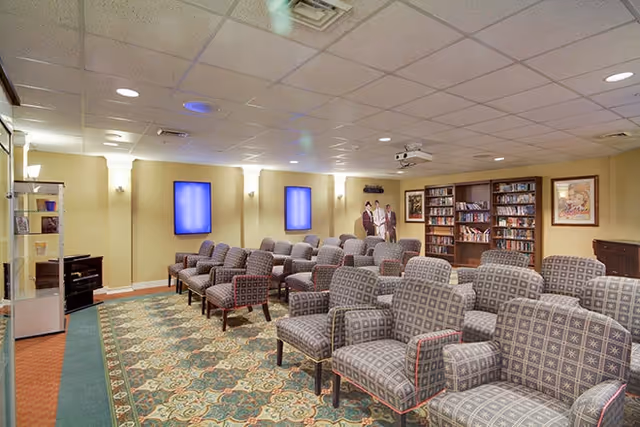 Carpeted community media room with rows of patterned armchairs facing a projector, bookshelves, and wall-mounted screens.
