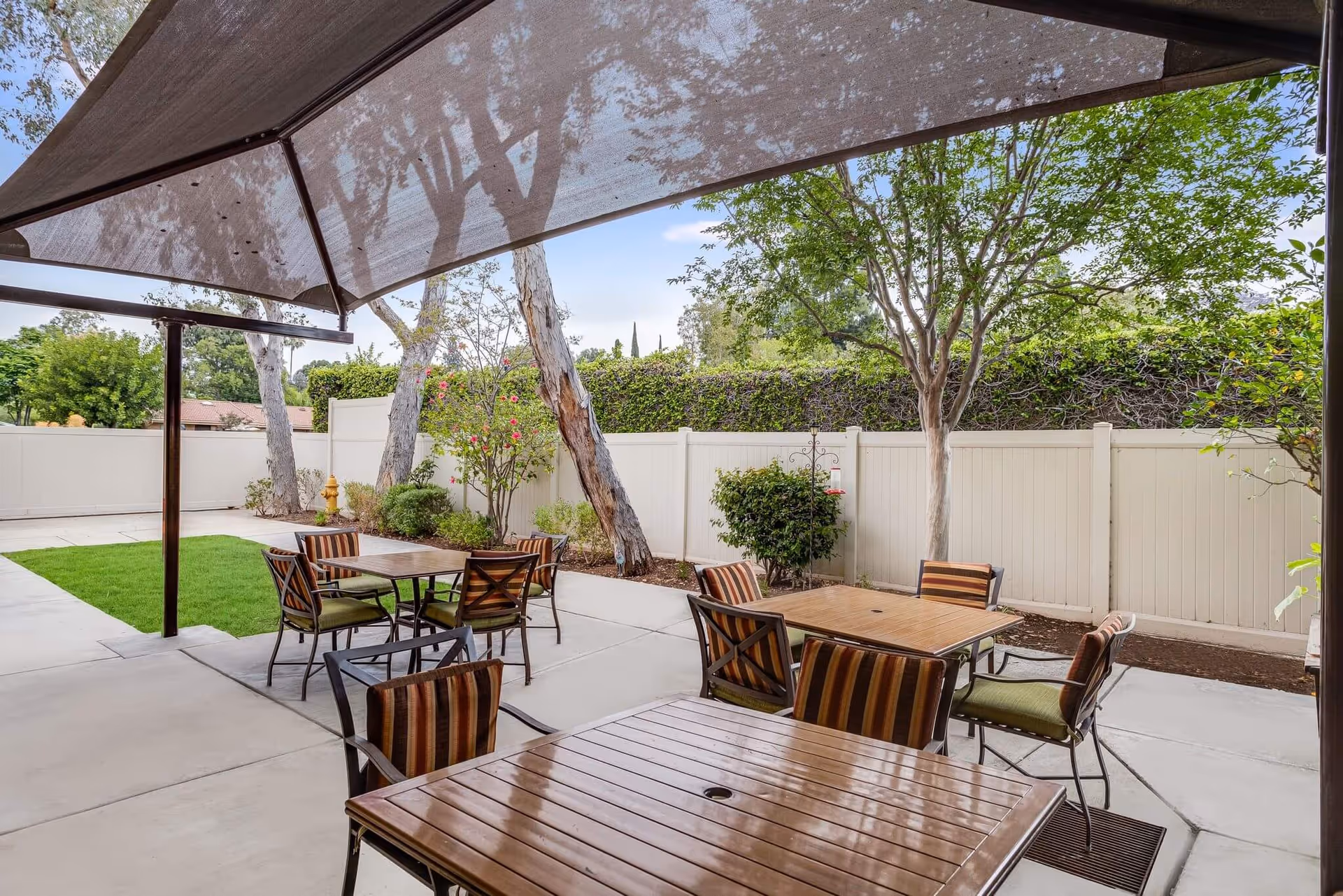 Outdoor patio area with several wooden tables and chairs with striped cushions under a large shade canopy. The patio is surrounded by a white fence and landscaped with trees, bushes, and grass patches.