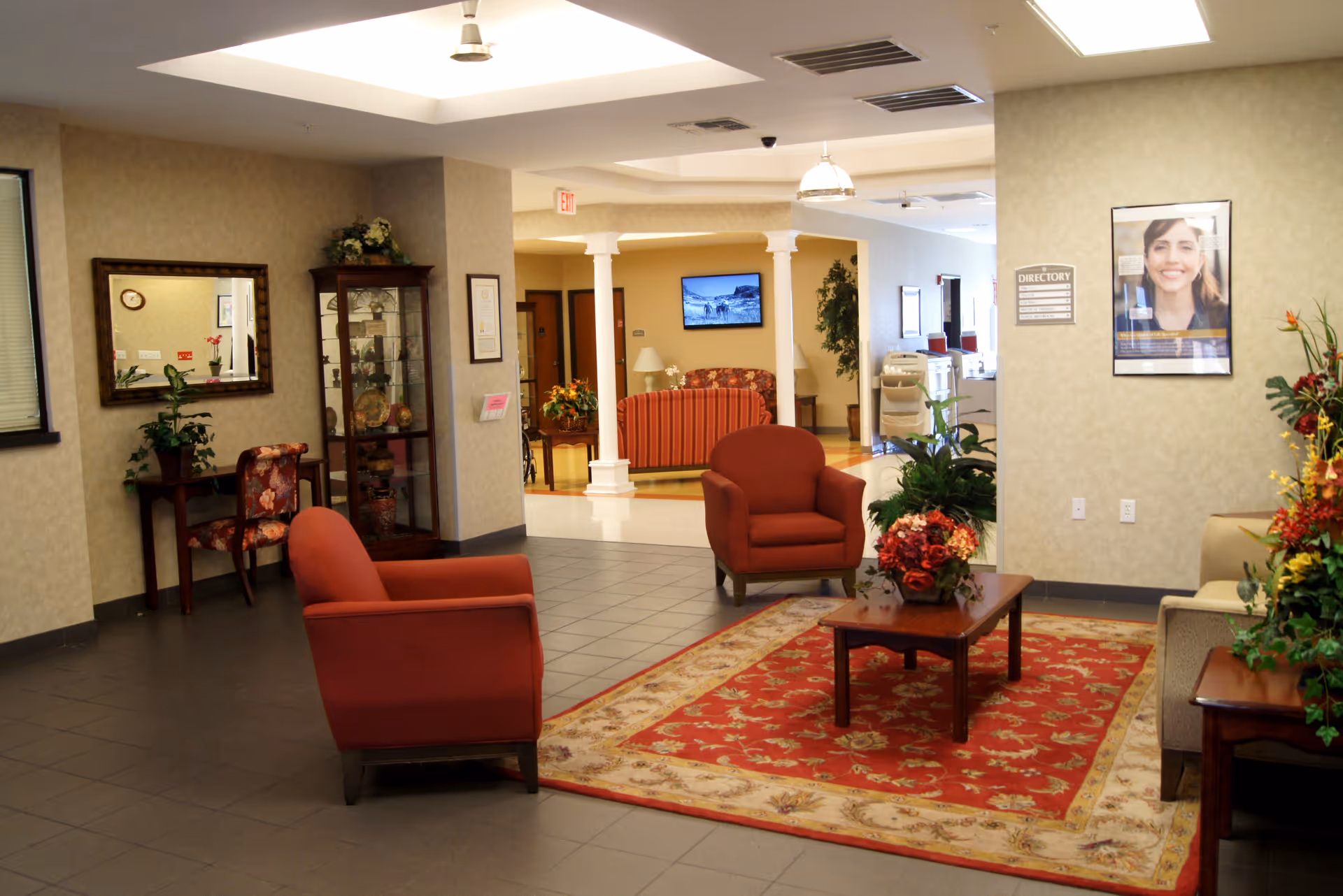 A nursing home lobby with red armchairs around a coffee table on a patterned rug, plants and a display cabinet with a reception area visible in the background.