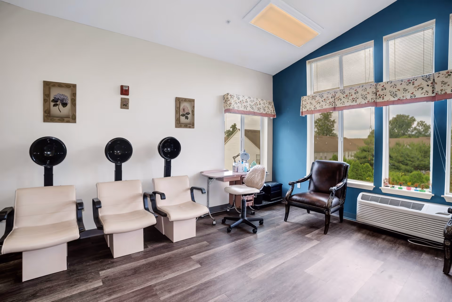 A bright salon room with three beige salon chairs equipped with black hair dryers, a small pink manicure table with a rolling chair, a dark brown leather armchair, and large windows with floral valances letting in natural light. The walls are painted white and blue, and the floor is dark wood.