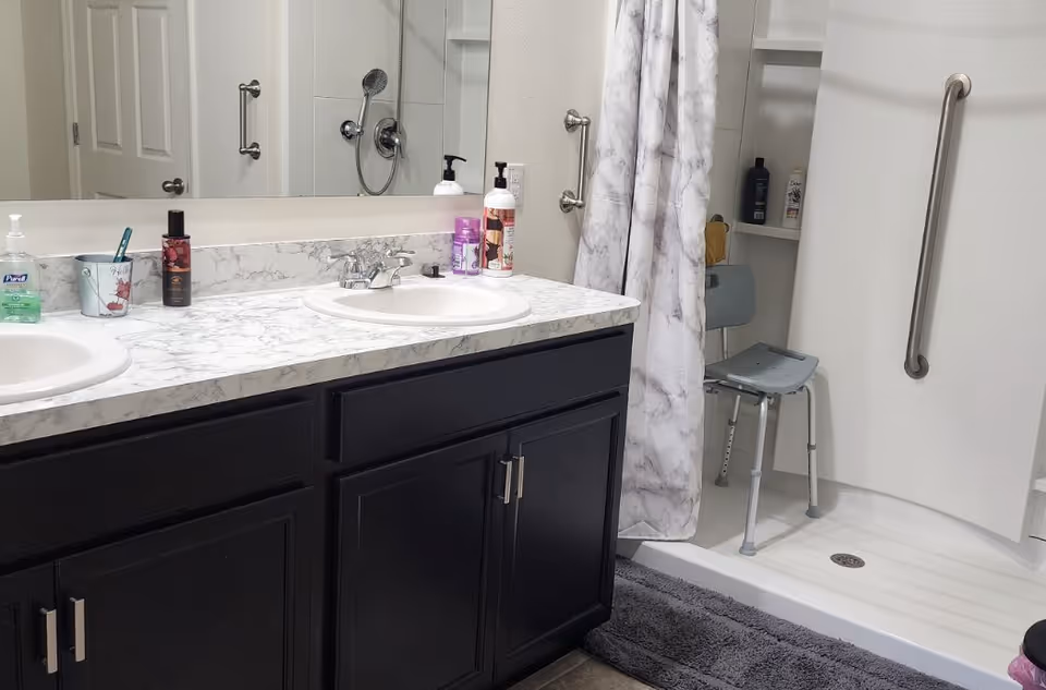 Accessible bathroom featuring a double-sink marble countertop over dark cabinets and a walk-in shower with a shower chair and grab bars.