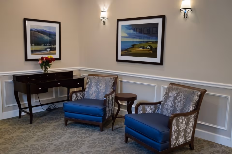 Two upholstered armchairs and a small side table beside a desk under framed landscape paintings in a sitting area.