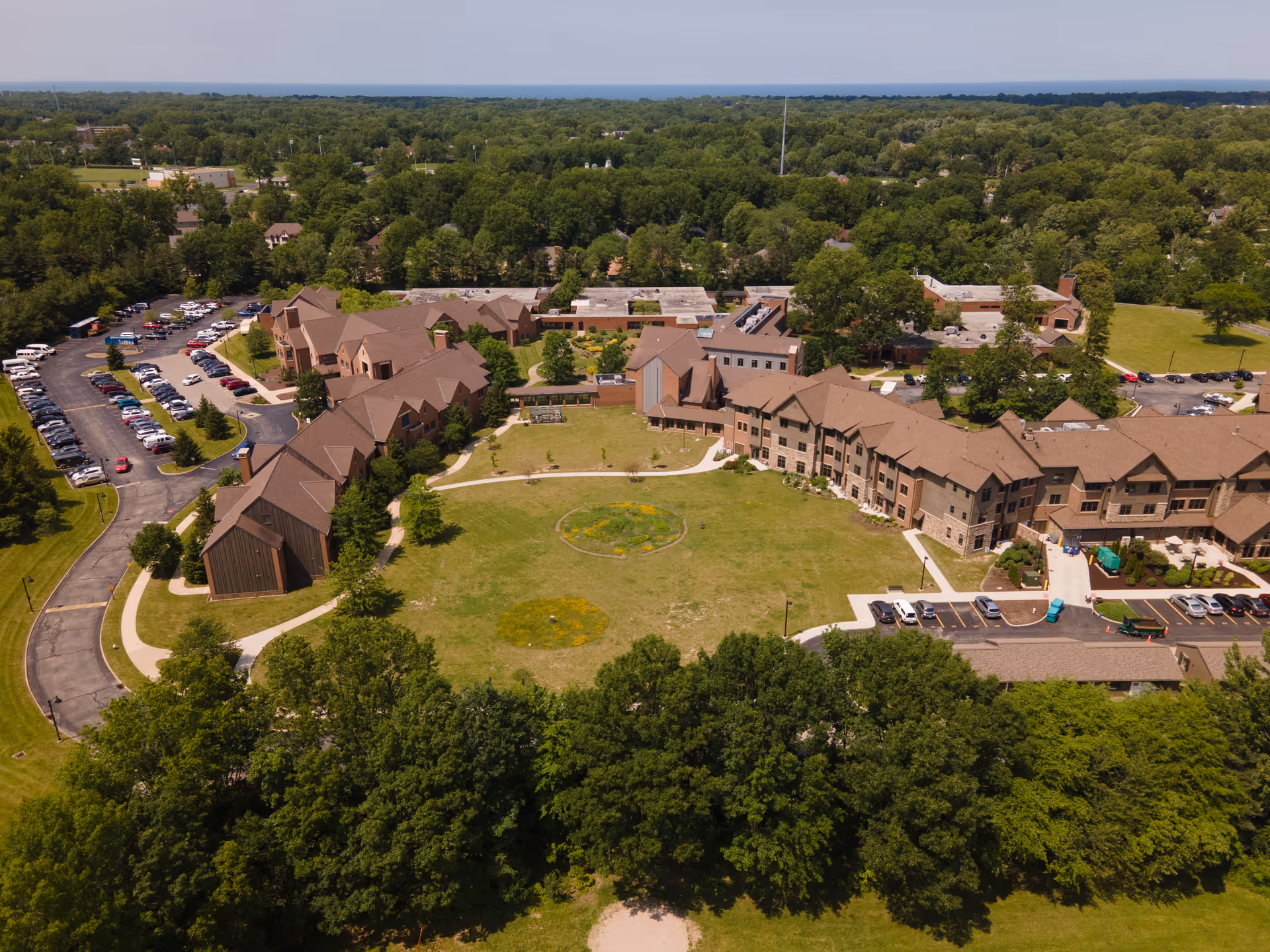 Aerial view of a senior living facility with brown-roofed buildings arranged around a large central lawn, parking areas, and surrounding trees.