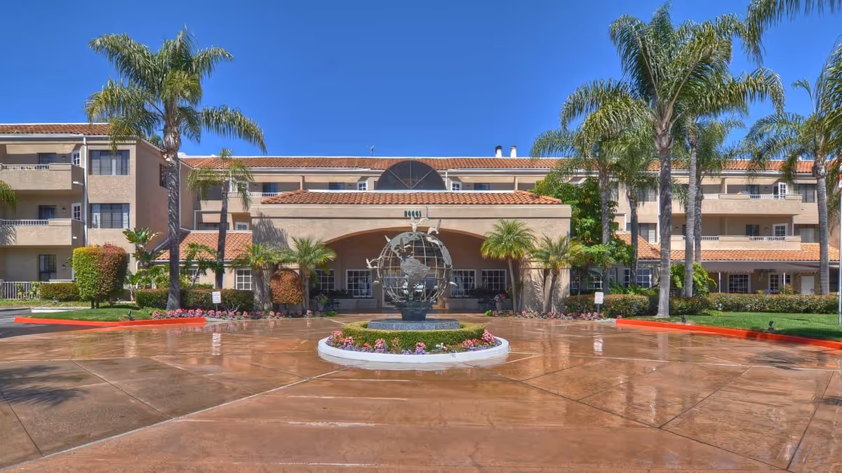 Front exterior view of Ivy Park at Laguna Woods, showing a three-story building with balconies, palm trees, and a circular driveway with a globe sculpture in the center surrounded by flowers.