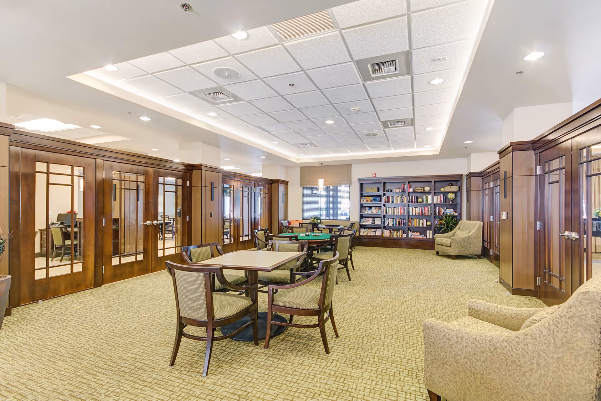 A spacious, well-lit common area with multiple tables and chairs arranged for social or recreational activities. The room features wood-paneled walls with glass doors, a carpeted floor, a bookshelf filled with books and decorative items, and comfortable armchairs. The ceiling has recessed lighting and a modern design.