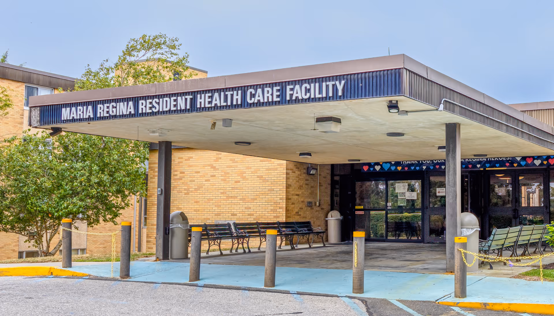 Front entrance canopy of the Maria Regina health care facility with benches, bollards, and glass doors.