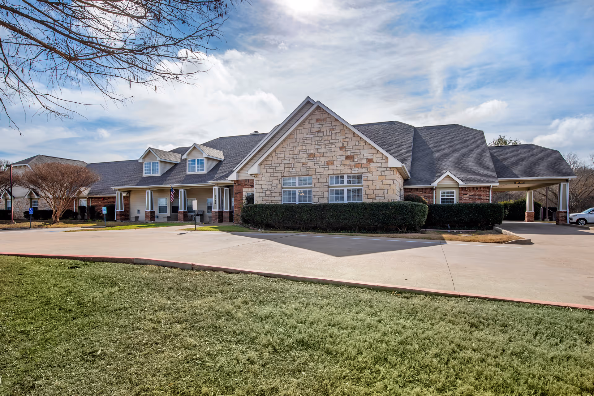 Exterior view of a single-story senior living facility building with a stone and brick facade, multiple windows, a covered entrance, and a driveway surrounded by grass and trees under a partly cloudy sky.