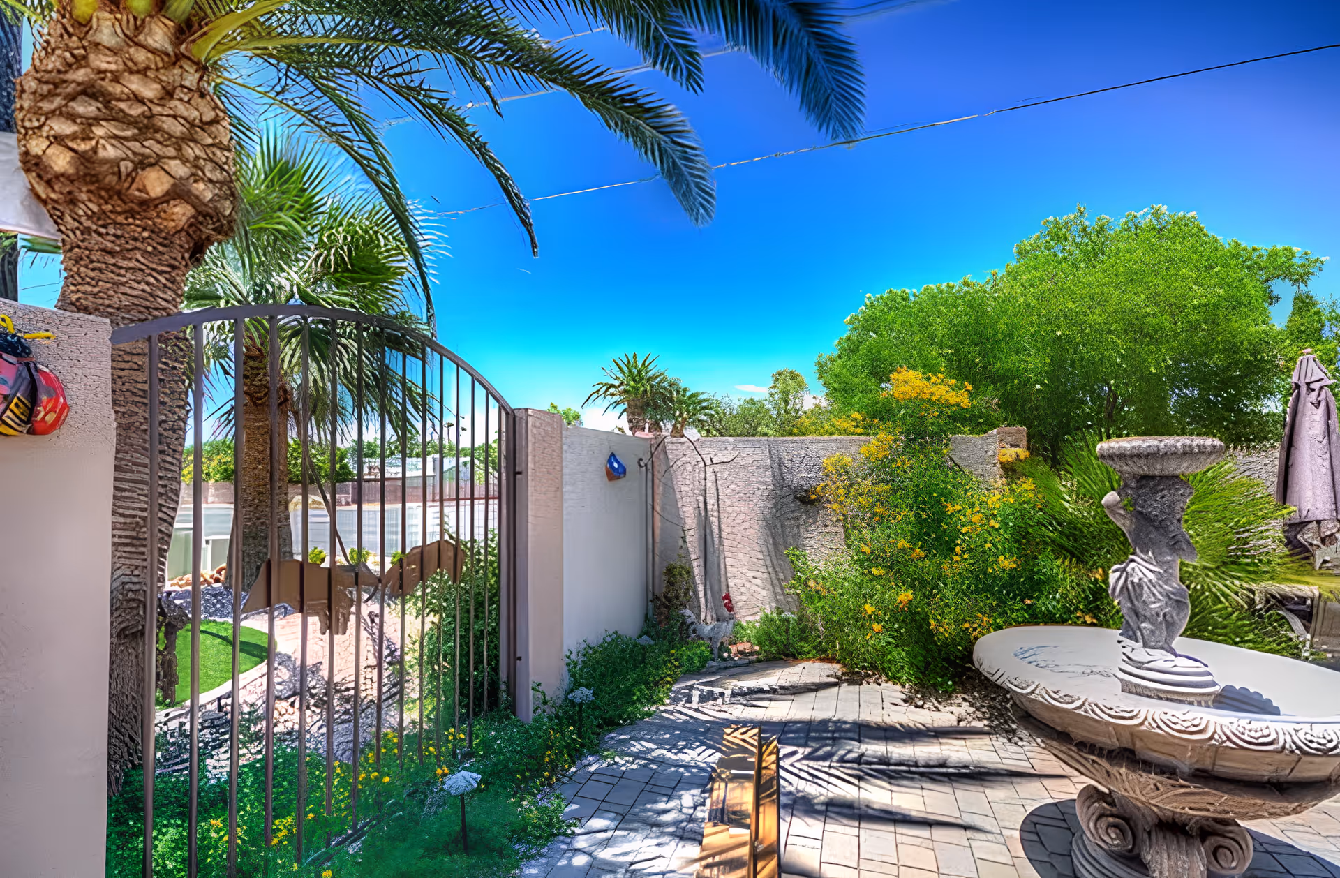Outdoor garden area with a stone birdbath on a pedestal, surrounded by lush green plants and yellow flowers. A metal gate with a decorative element is visible on the left side, along with palm trees and a clear blue sky overhead. The ground is paved with stone tiles, and there is a folded umbrella in the background.