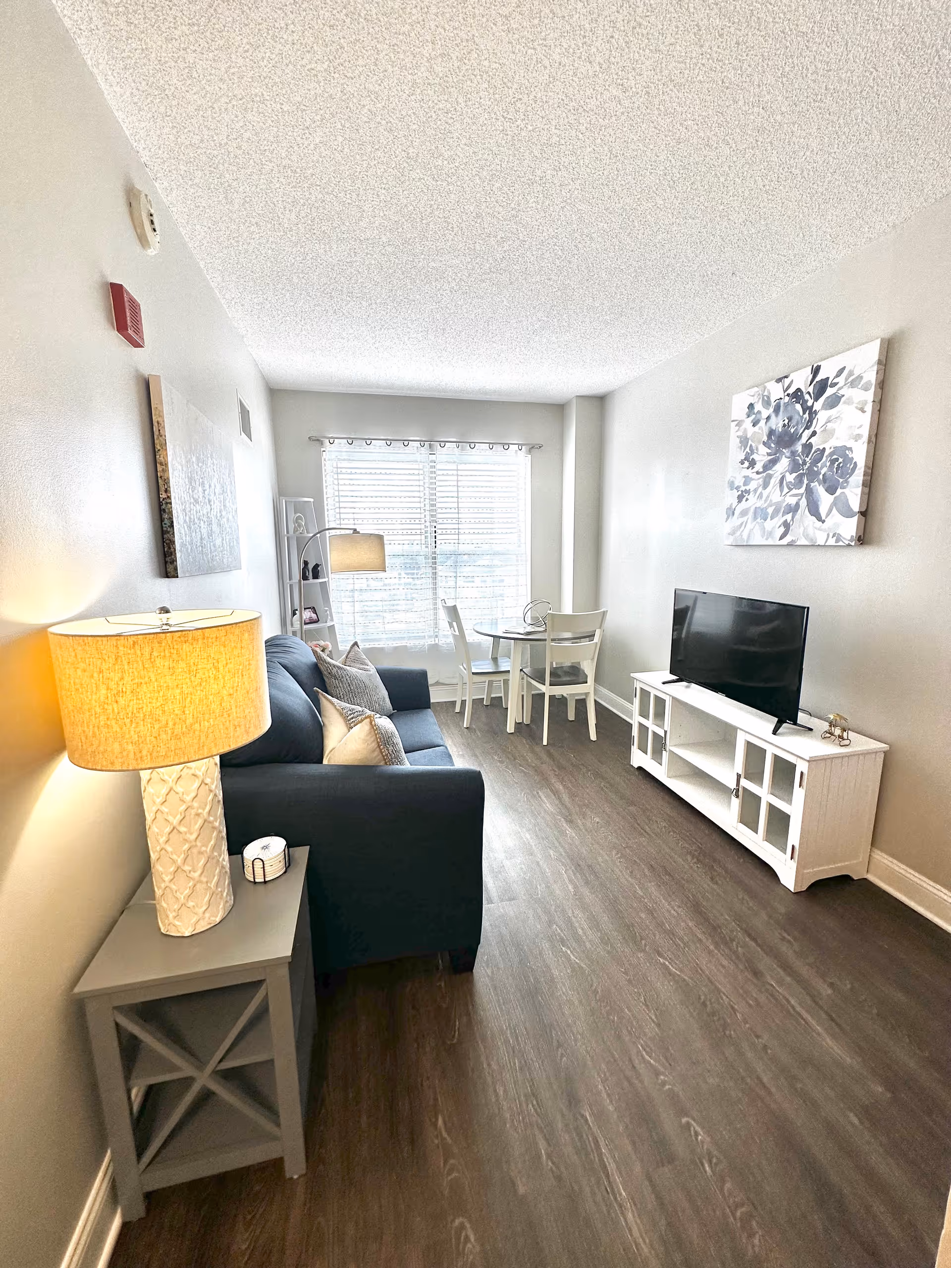 A bright living room with a dark blue sofa adorned with pillows, a side table with a lit lamp, a white TV stand with a flat-screen television, a small round dining table with two chairs near a window with blinds, and a floral painting on the wall.