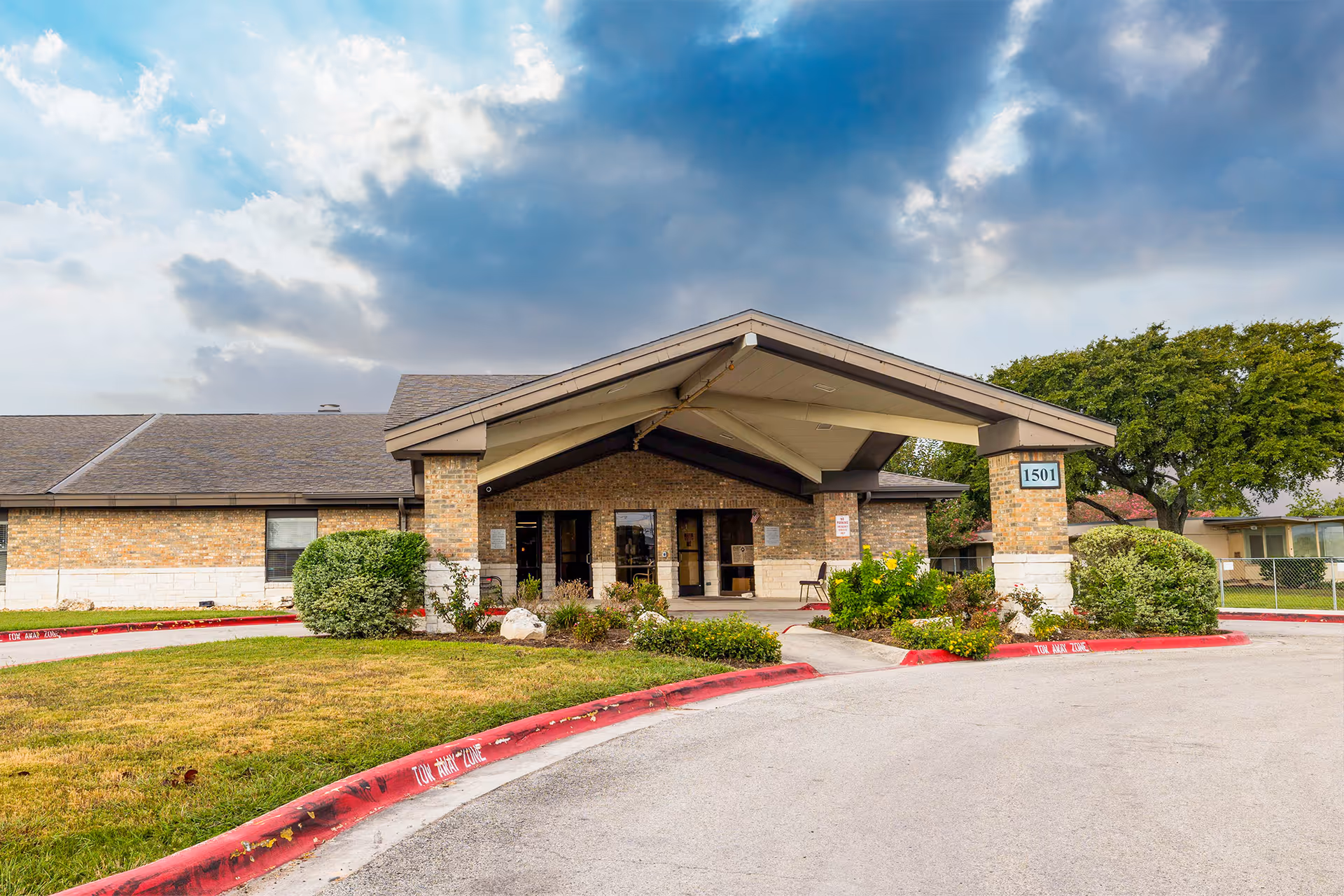 Front exterior view of Parkview Nursing and Rehabilitation Center showing a single-story brick building with a covered entrance, surrounded by bushes and a curved driveway with red curbs under a partly cloudy sky.