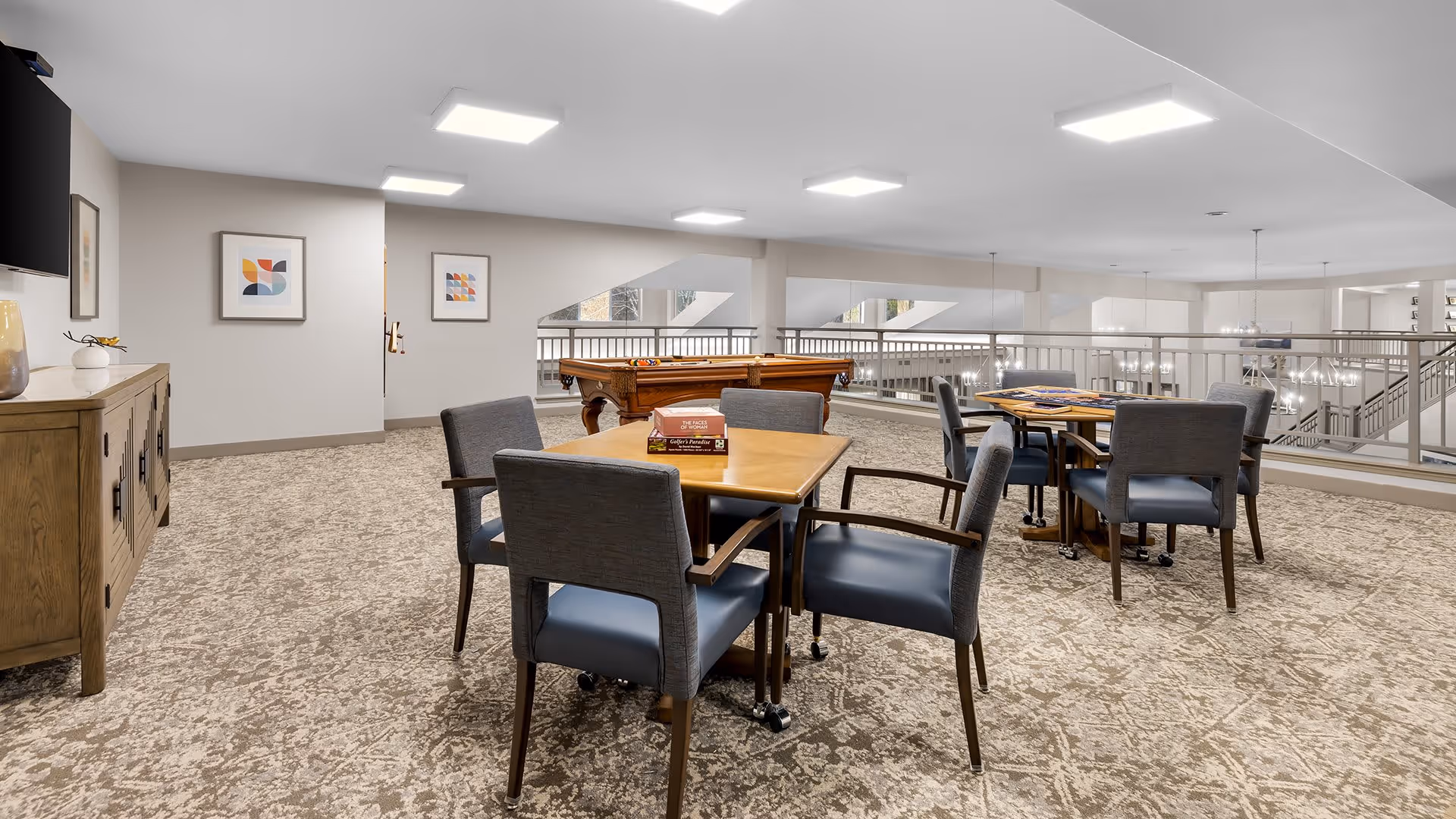A spacious common area in a senior living facility featuring two wooden tables with gray cushioned chairs around them. There are board games on the tables. In the background, there is a pool table and a wooden sideboard with decorative items. The room has carpeted flooring, white walls with framed abstract art, and bright ceiling lights. A railing overlooks a lower level with additional lighting fixtures.