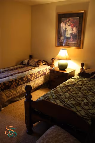 A cozy bedroom with two single beds separated by a wooden nightstand. The nightstand has a green lamp with a beige lampshade. Above the nightstand hangs a framed floral painting. The beds have patterned bedspreads in shades of brown and green.