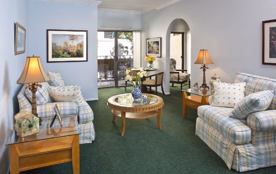 Well-appointed living room with two plaid sofas, a round glass-top coffee table, side tables with lamps, and a sliding glass door to a balcony.