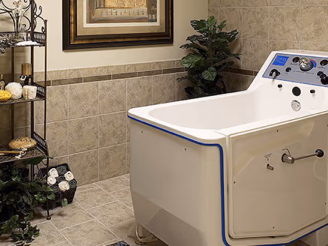 A tiled bathroom with a large white medical bathtub featuring control knobs and a digital display. The room has beige tiled walls and floor, a metal rack with towels and bath items, a green plant in the corner, and a framed picture on the wall.