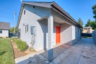 Exterior view of a single-story building with light gray walls and a bright red door under a covered porch area. The building is surrounded by a concrete walkway and a small grassy area with some plants. Other similar buildings and trees are visible in the background under a clear blue sky.